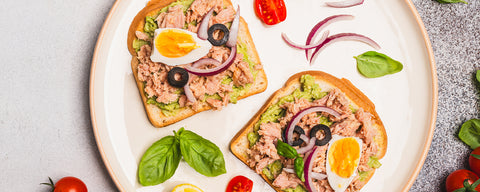 Two avocado toasts topped with tuna, boiled egg, red onion, and black olives on a white plate with basil and cherry tomatoes.
